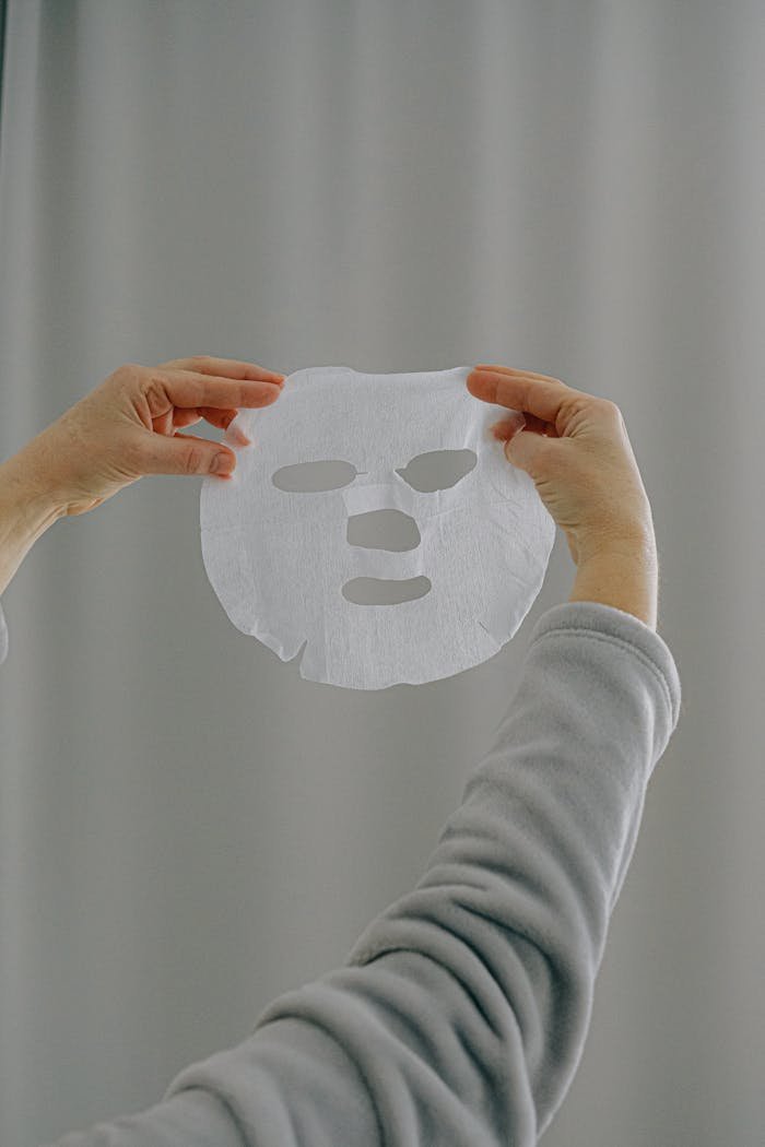 Hands presenting a white sheet mask against a neutral background, promoting skincare and wellness.